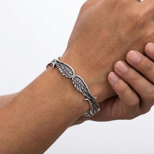 K&ouml;nnte beinhalten: Ein silberfarbenes Armband mit detailliertem Fl&uuml;geldesign. Das Armband umschlie&szlig;t das Handgelenk, wobei sich die Fl&uuml;gel nach au&szlig;en erstrecken. Die filigrane Federung verleiht diesem Schmuckst&uuml;ck eine einzigartige Note.