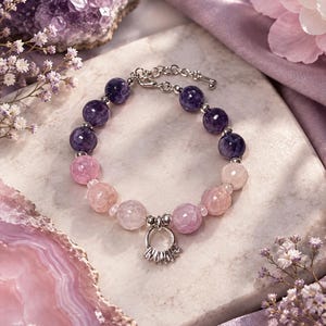 Op de afbeelding: Een kralenarmband met gefacetteerde donkerpaarse amethist en lichtroze rozenkwarts kralen in een kleurverloop. De armband is voorzien van een zilverkleurige metalen bedel in de vorm van een kleine ring met kleine doorzichtige steentjes en een zilverkleurige sluiting.