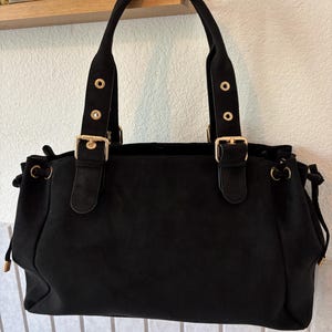 Peut inclure: Un sac à main noir à la texture mate et douce, doté de deux anses fixées par des boucles et des œillets en métal doré. Les côtés du sac présentent des détails de cordon de serrage avec des embouts en métal doré, offrant une forme structurée.