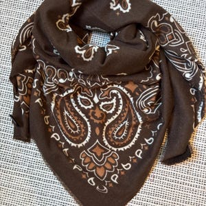 Peut inclure: Un foulard triangulaire marron foncé orné d'un motif cachemire blanc et marron clair. Le tissu présente une texture douce avec des bords effilochés sur les côtés. Le design inclut des motifs floraux et en forme de goutte dans un style bandana.