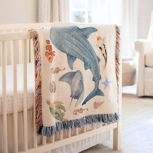 Könnte beinhalten: Eine beige gewebte Decke mit Illustrationen von Meereslebewesen, die über einem Kinderbett drapiert ist. Das Design umfasst einen Walhai, einen Stechrochen, einen Clownfisch, ein Seepferdchen, eine Schildkröte und einen Seestern. Die Decke hat einen Fransenrand.