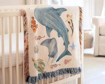 May include: A woven throw blanket featuring a cream-colored background with a large blue whale shark, a stingray, a sea turtle, a clownfish, a koi fish, a starfish, a jellyfish, and two seahorses. The blanket has decorative multi-colored fringe edges.