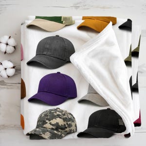 Peut inclure: Une couverture en polaire blanche ornée d'un motif répétitif de casquettes de baseball dans des tons gris anthracite, violet, jaune moutarde, vert olive et camouflage, avec un coin replié révélant un envers blanc et doux.