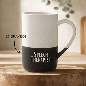Puede incluir: Una taza de cerámica de dos tonos con acabado moteado. La mitad superior es de color blanco roto y la inferior es negra. El texto "SPEECH THERAPIST" está grabado en blanco en la sección negra. Una flecha señala el texto con la etiqueta "ENGRAVED!".
