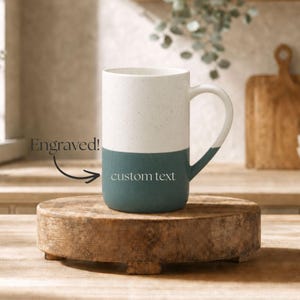 May include: A two-toned ceramic mug with a speckled white upper section and a solid teal lower section. The mug features a handle on the right side. The text "Engraved!" is visible with an arrow pointing to the teal area labelled "custom text".