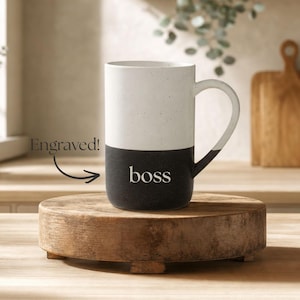 May include: A two-toned ceramic mug with a speckled white upper section and a solid black lower section. The word "boss" is engraved in white on the black portion. A curved handle extends from the right side of the mug, matching the black colour.