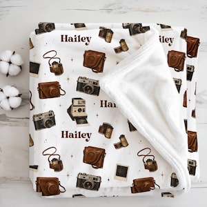 May include: A white fleece blanket featuring a repeating pattern of brown vintage cameras, leather camera cases, film rolls, and blank photo frames, interspersed with small gold stars and the name Hailey printed in a dark brown serif font.