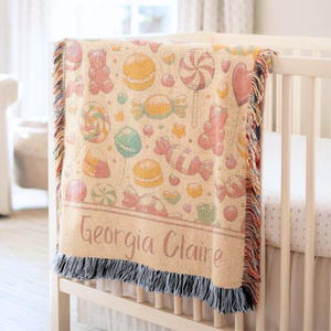 Puede incluir: Una manta de bebé beige con un estampado de caramelos, que incluye piruletas, ositos de goma y macarons en colores pastel. La manta tiene un borde con flecos y el nombre "Georgia Claire" está impreso.