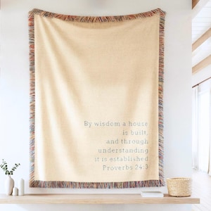 Pode incluir: Um cobertor tecido bege com bordas franjadas, exibindo o texto "By wisdom a house is built, and through understanding it is established Proverbs 24:3." O cobertor está pendurado em uma parede branca.