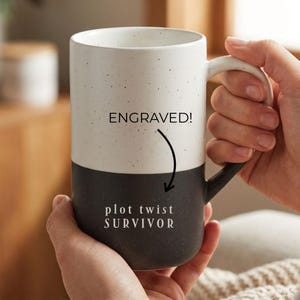May include: A two-toned ceramic mug with a speckled finish, featuring a white upper section and a black lower section. The text ENGRAVED! appears above a curved arrow pointing to the text plot twist SURVIVOR printed on the black base.