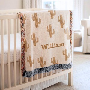 Könnte beinhalten: Eine gewebte Wohndecke mit einem sich wiederholenden Muster aus beigen Kaktussilhouetten auf cremefarbenem Grund. Der Name William ist mittig in beigem Text eingestickt. Die Decke hat an den Seiten und am unteren Rand mehrfarbige Fransen.