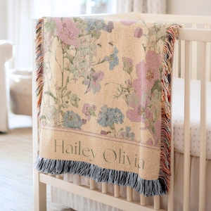 Könnte beinhalten: Eine beige Blumendecke mit einem botanischen Design in Rosa-, Blau- und Grüntönen, drapiert über einem weißen Kinderbett. Die Decke hat einen Fransenrand und der Name "Hailey Olivia" ist unten abgebildet.