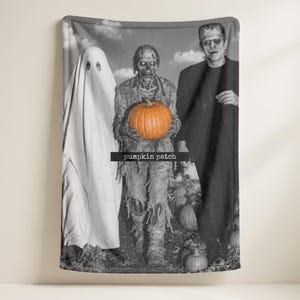 May include: A black and white throw blanket featuring a ghost, a zombie holding a bright orange pumpkin, and a man in a suit. The text "pumpkin patch" is printed in a black rectangular bar across the centre of the blanket design.