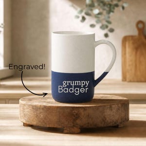 May include: A two-toned ceramic mug with a speckled white upper section and a solid dark blue lower section. The text "grumpy Badger" is engraved in white on the blue portion. An arrow points to the text with the label "Engraved!"