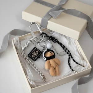 Könnte beinhalten: Ein Schlüsselanhänger-Set mit einem kleinen beigen Teddybären, der einen weiß-schwarzen Helm trägt, einem schwarzen Anhänger mit dem Text Holy Bible, einem weißen Golfball und einem silbernen Karabiner sowie zwei schwarz-weiß gemusterten Kordeln.