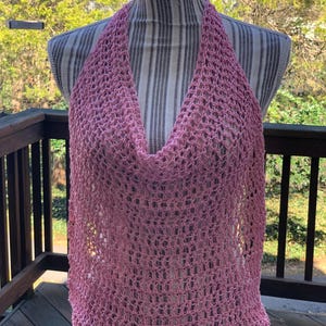 Può includere: Top all'uncinetto rosa chiaro, esposto su un manichino. Il top presenta uno scollo a cascata e un design ampio e arioso. Gli spallacci sono larghi e il corpo del top ha un aspetto a rete. Lo sfondo mostra un ponte di legno e vegetazione.