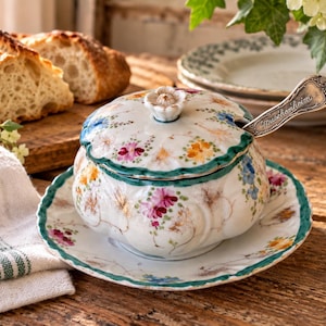 May include: A white porcelain sugar bowl with a matching saucer, featuring hand-painted floral motifs in pink, yellow, and blue, with a green scalloped rim. A silver spoon with the engraved text "Meissenheim" rests inside the lidded bowl.