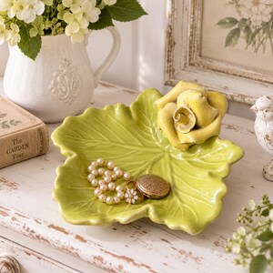 May include: A lime green ceramic trinket dish shaped like a large leaf with a sculpted yellow rose attached to one side. The dish contains a strand of white faux pearls, a gold-toned flower ring, and a small round gold-toned metal locket.