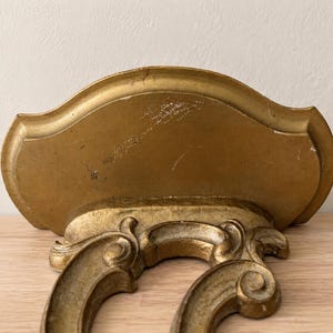May include: A gold-colored wall shelf featuring a curved, semi-circular top surface and an ornate, scrolled bracket support underneath. The finish shows signs of wear and minor surface scratches, with a metallic gold paint application throughout.