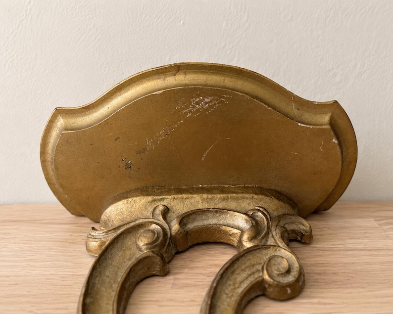 May include: A gold-colored wall shelf featuring a curved, semi-circular top surface and an ornate, scrolled bracket support underneath. The finish shows signs of wear and minor surface scratches, with a metallic gold paint application throughout.