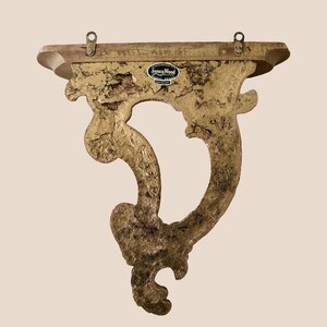 May include: A gold-colored wall shelf with a textured, ornate bracket featuring a curved, scroll-like design. The top shelf is flat and rectangular with two metal mounting loops attached to the back. A black label reads Syroco Wood.