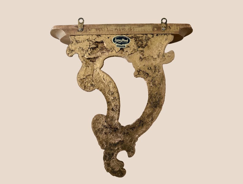 May include: A gold-colored wall shelf with a textured, ornate bracket featuring a curved, scroll-like design. The top shelf is flat and rectangular with two metal mounting loops attached to the back. A black label reads Syroco Wood.