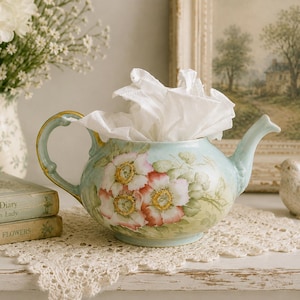 May include: A light blue ceramic teapot with a hand-painted floral design featuring pink and white blossoms with yellow centers. The teapot has a curved handle with gold trim and a spout, and it is filled with white tissue paper.