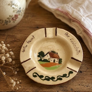 May include: A round, cream-colored ceramic ashtray featuring a hand-painted illustration of a small house with a red roof and green trees. The rim displays the text "who burnt the tablecloth" in black script, accented by brown lines.
