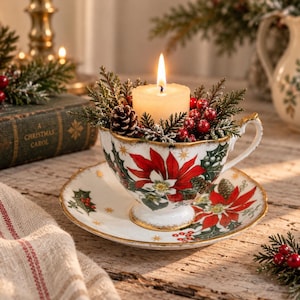 May include: A white porcelain teacup and saucer set decorated with red poinsettia flowers, green holly leaves, and gold accents. A lit cream-colored pillar candle sits inside the cup, surrounded by small pinecones, evergreen sprigs, and red berries.