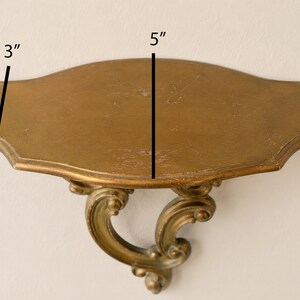 May include: A gold-colored wall shelf with a curved, ornate support bracket. The shelf surface is oval-shaped with scalloped edges. Text on the image indicates a depth of 3 inches and a width of 5 inches, providing clear physical dimensions.