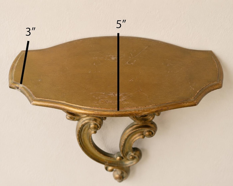 May include: A gold-colored wall shelf with a curved, ornate support bracket. The shelf surface is oval-shaped with scalloped edges. Text on the image indicates a depth of 3 inches and a width of 5 inches, providing clear physical dimensions.