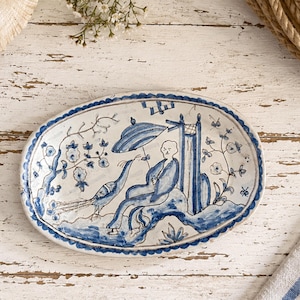 May include: An oval ceramic dish featuring a hand-painted blue and white illustration of a person sitting under an umbrella next to a peacock. The design includes floral motifs and a decorative scalloped border, all rendered in a classic blue pigment.