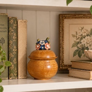 May include: A round, amber-colored ceramic jar with a lid featuring a sculpted cluster of small, multi-colored flowers in shades of pink, blue, and white. The jar has a smooth, glazed finish and a rounded body, sitting on a flat base.