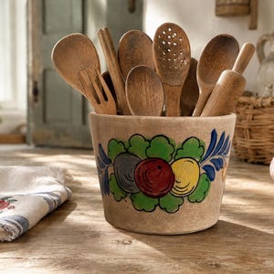 May include: A beige ceramic crock featuring a hand-painted design of green leaves and three circular fruits in red, yellow, and blue. The crock is filled with an assortment of wooden kitchen utensils, including spoons, a fork, and a rolling pin.