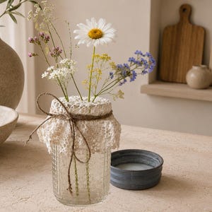 May include: A clear glass jar with a textured grid pattern, adorned with a cream-colored crocheted lace cover and a thin brown twine bow. The jar contains a small bouquet of wildflowers, including a white daisy, yellow sprigs, and blue flowers.