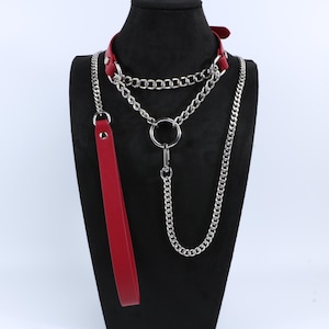 May include: A red leather collar with a silver chain and a matching leash. The collar has a buckle closure and multiple silver chain strands. The leash is a long, thin strip of red leather.