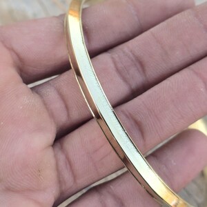 Può includere: Un braccialetto rigido color oro con un profilo piatto e rettangolare. Il braccialetto è liscio e lucido, riflettendo la luce. Il gioiello è un design semplice ed elegante.