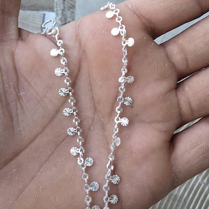 May include: A delicate silver anklet featuring a chain adorned with small, circular and starburst-shaped charms. The anklet is displayed against a neutral background, showcasing its intricate design and reflective qualities. This piece of jewelry is designed to be worn around the ankle.