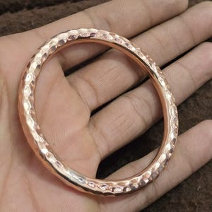 Op de afbeelding: Een gehamerde koperen armband met een getextureerd oppervlak. De armband is een massieve band van koper met een cirkelvorm. Het koper heeft een warme, roodbruine kleur en een glanzende afwerking. De armband wordt in een hand gehouden.