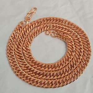 Può includere: Una collana a catena color rame con un design a maglie a catena. La collana è realizzata in metallo e presenta una chiusura a moschettone. La catena è arrotolata e poggia su una superficie bianca. Le maglie sono spesse e strettamente collegate.