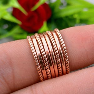 Op de afbeelding: Een verzameling roségouden ringen, sommige met een gladde afwerking en andere met een gedraaid touwontwerp. De ringen zijn op een vinger gestapeld en tonen hun verschillende texturen en stijlen. De achtergrond toont een wazige rode bloem en groen gebladerte.