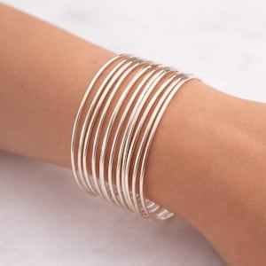 May include: A stack of silver bangles on a wrist. The bangles are thin, round, and shiny, reflecting light. The jewelry is worn on a person's arm, showcasing the minimalist design. The bracelets are a simple, elegant accessory.