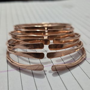 May include: A stack of copper-colored metal bracelets. Each bracelet has a hammered texture and an open design. The bracelets are stacked on top of each other, showcasing their individual shapes and the warm tones of the metal.