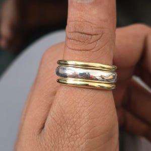 Puede incluir: Un anillo de tres bandas con una banda central plateada y bandas exteriores doradas. El anillo está en un dedo, mostrando el contraste entre los metales. Las bandas tienen un acabado liso y pulido, que refleja la luz.