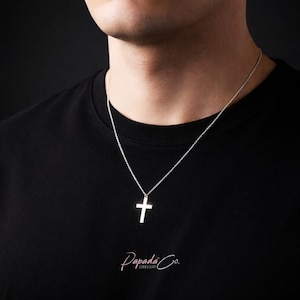 Op de afbeelding: Een zilveren kruis-hanger ketting. Het kruis heeft een eenvoudig ontwerp en hangt aan een fijne ketting. De ketting wordt gedragen op een zwart T-shirt. Het Papada Co. Jewellery logo is onderaan zichtbaar.