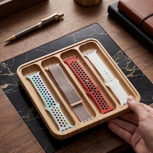 Puede incluir: Una bandeja de madera con cuatro correas de reloj en varios colores: azul claro, beige, rojo y blanco. Las correas están en compartimentos separados. Un bolígrafo dorado descansa sobre la superficie. Una mano sostiene la bandeja.