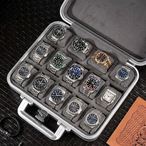 Pode incluir: Uma mala prateada aberta, exibindo uma coleção de relógios de pulso de luxo. Os relógios apresentam vários designs, incluindo mostradores pretos, azuis e dourados, com pulseiras prateadas e douradas. A mala é forrada com tecido cinza.