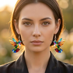 May include: A pair of dangling earrings featuring colorful hummingbird pendants. Each bird has a stained-glass appearance with vibrant segments of red, orange, yellow, green, and blue, suspended from simple gold-toned metal hooks.