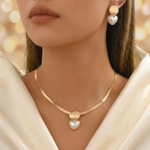 Peut inclure: Ensemble de bijoux dorés comprenant un collier et des boucles d'oreilles. Le collier est composé d'une chaîne en chevrons avec un pendentif en forme de disque doré et d'une perle en forme de cœur. Les boucles d'oreilles ont un design similaire.