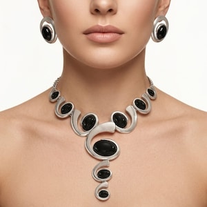 Puede incluir: Un conjunto de collar y pendientes plateados con piedras negras ovaladas. El collar presenta un dise&ntilde;o &uacute;nico y fluido con elementos curvos. Los pendientes son ovalados con una piedra negra en el centro, complementando el estilo del collar.